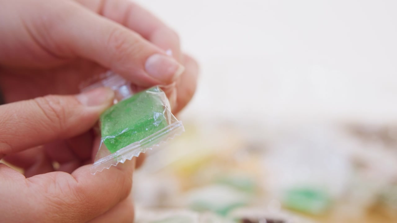 Closeup hands unwrapping green candy, transparent wrapper peeled to reveal sugar crystals, focused fingertips and careful peeling motion, blurred colorful candies in background, indoor countertop