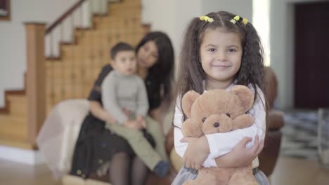 sonriente chica de oriente medio con ojos marrones y cabello rizado sosteniendo el oso de peluche y mirando a la cámara