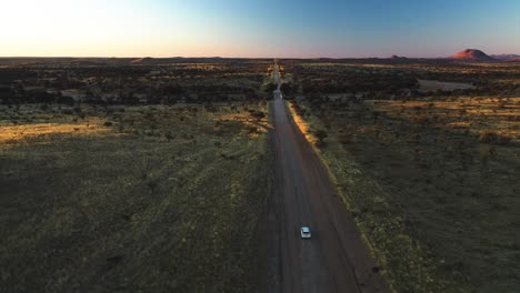 White-SUV-driving-on-a-dirt-road-through-wastelands-of-Namibia,-sundown-in-Africa---Aerial-view