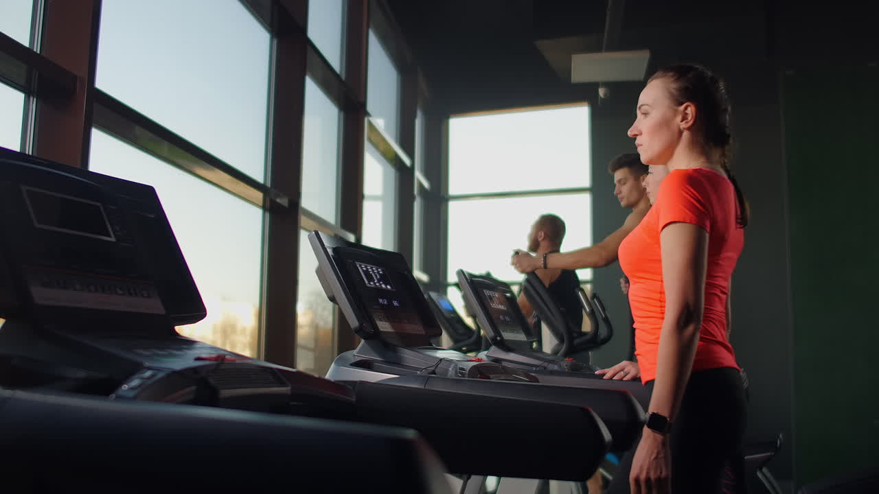A young beautiful girl walks on a treadmill in front of a large panoramic window in slow motion against the background of several people.
