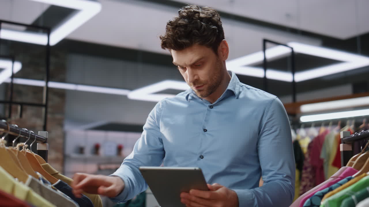 trabajador de una boutique caminando en filas de ropa comprobando la tableta en una tienda de ropa de primer plano