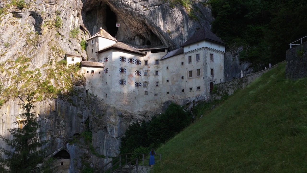Una turista si trova di fronte al Castello di Predjama, una fortezza medievale incastonata in una scogliera nella regione della Carniola Interna in Slovenia. Ripresa aerea in allontanamento.
