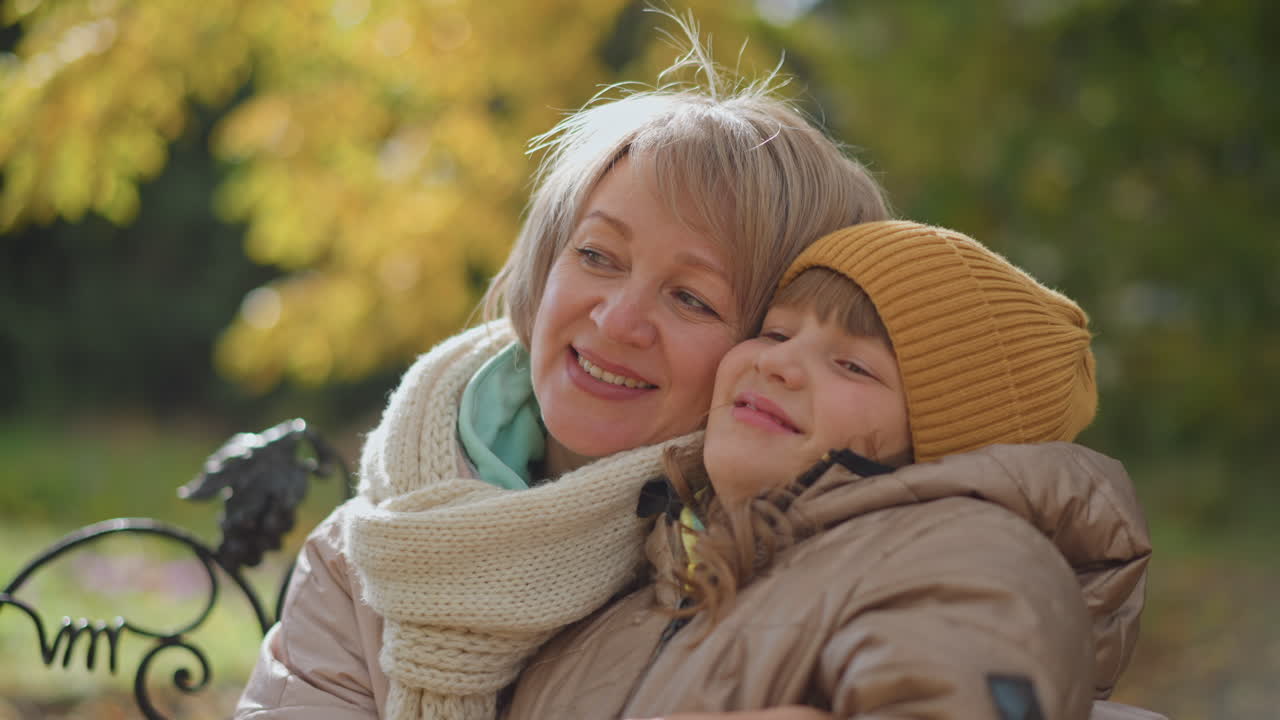 mother holds daughter affectionately on park bench as leaves sway gently in wind through autumn foliage, both smiling in pastel coats and knit scarf capturing tender family moment