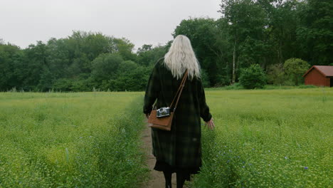 blonde cute german girl walking across a field of flowers in a black coat vintage bag and camera towards a forest on misty cloudy scandinavian spring touching with hands wanderlust exploring adventure