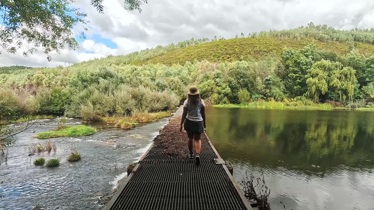 A girl hikes across a bridge over the Mondego River in Portugal, surrounded by lush nature and flowing water