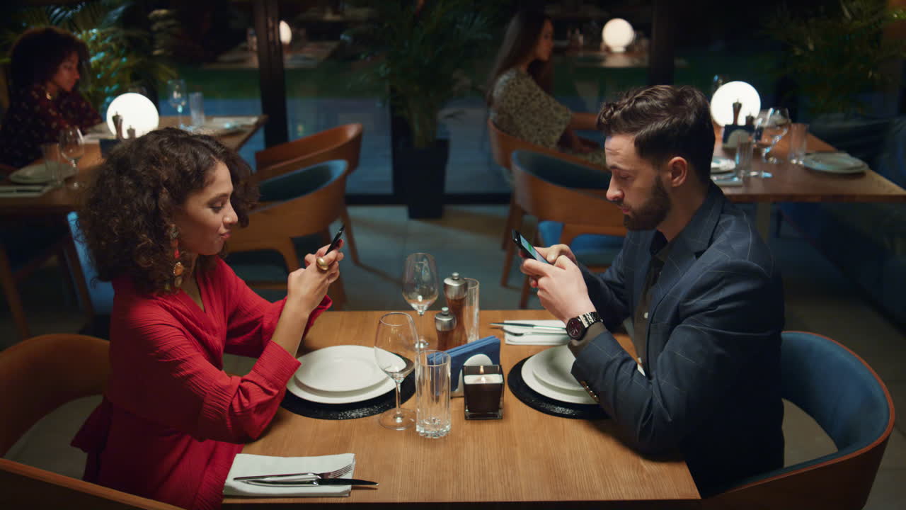 Partner multietnici che consultano il telefono durante un appuntamento a cena in un ristorante elegante.