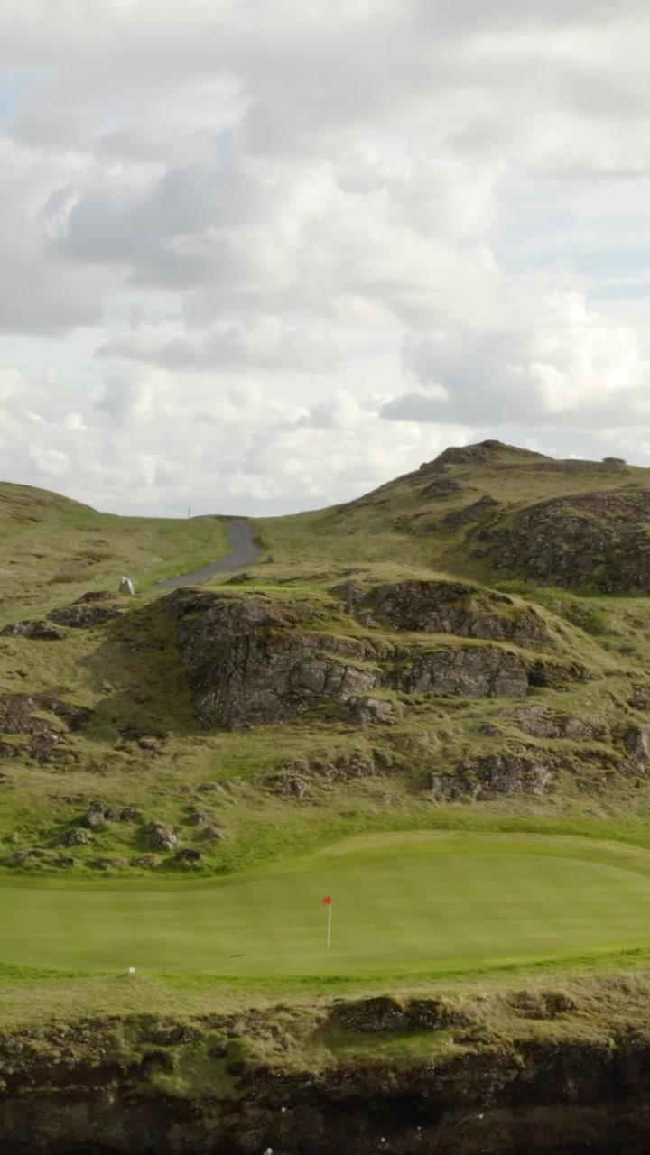Un vibrante green da putting di un campo da golf con una bandiera rossa si trova alla base di una montagna aspra e rocciosa sotto un cielo nuvoloso