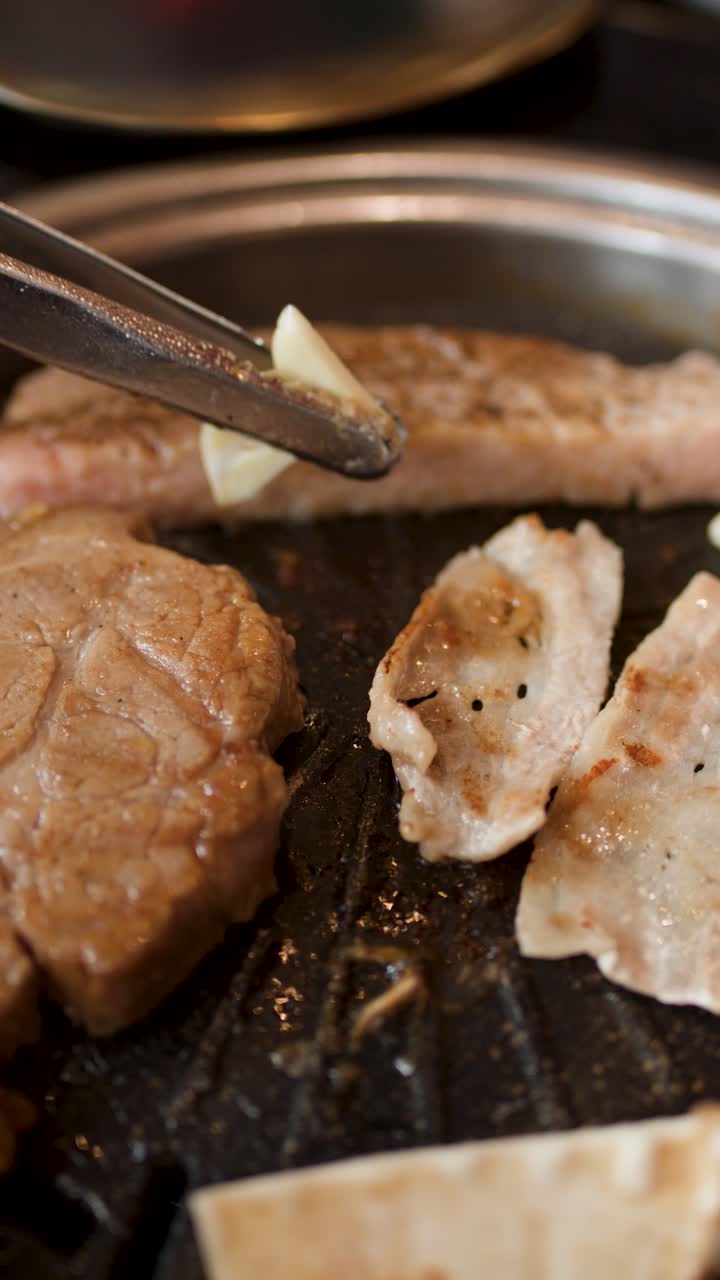 Tongs place garlic slice onto sizzling pork belly on tabletop Korean barbecue grill, warm lighting