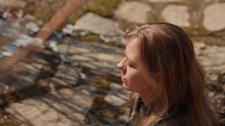 Giovane donna mostrata di profilo che cammina lungo un sentiero di pietra illuminato dal sole, con un'atmosfera calma e contemplativa, una leggera inclinazione in avanti, ciocche di capelli sollevate, sguardo abbassato e una luce naturale soffusa che illumina i dintorni tranquilli.
