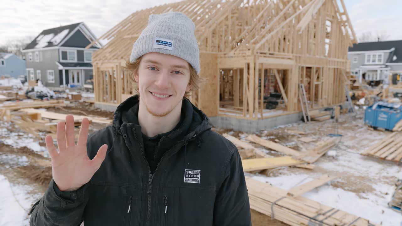 A Young Builder Waves in the Snowy Construction Site, Showcasing the Progress of a New Home with Wooden Frameworks Under a Cloudy Sky