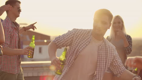 the young men is dancing on the roof with his friends who drinks beer on the party. he smiles and enjoys the time in shorts and a blule plaid shirts in summer evening.