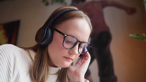 close up of young woman with glasses and headphones deeply focused while listening to music with blurred wall painting and warm ambient lights in background