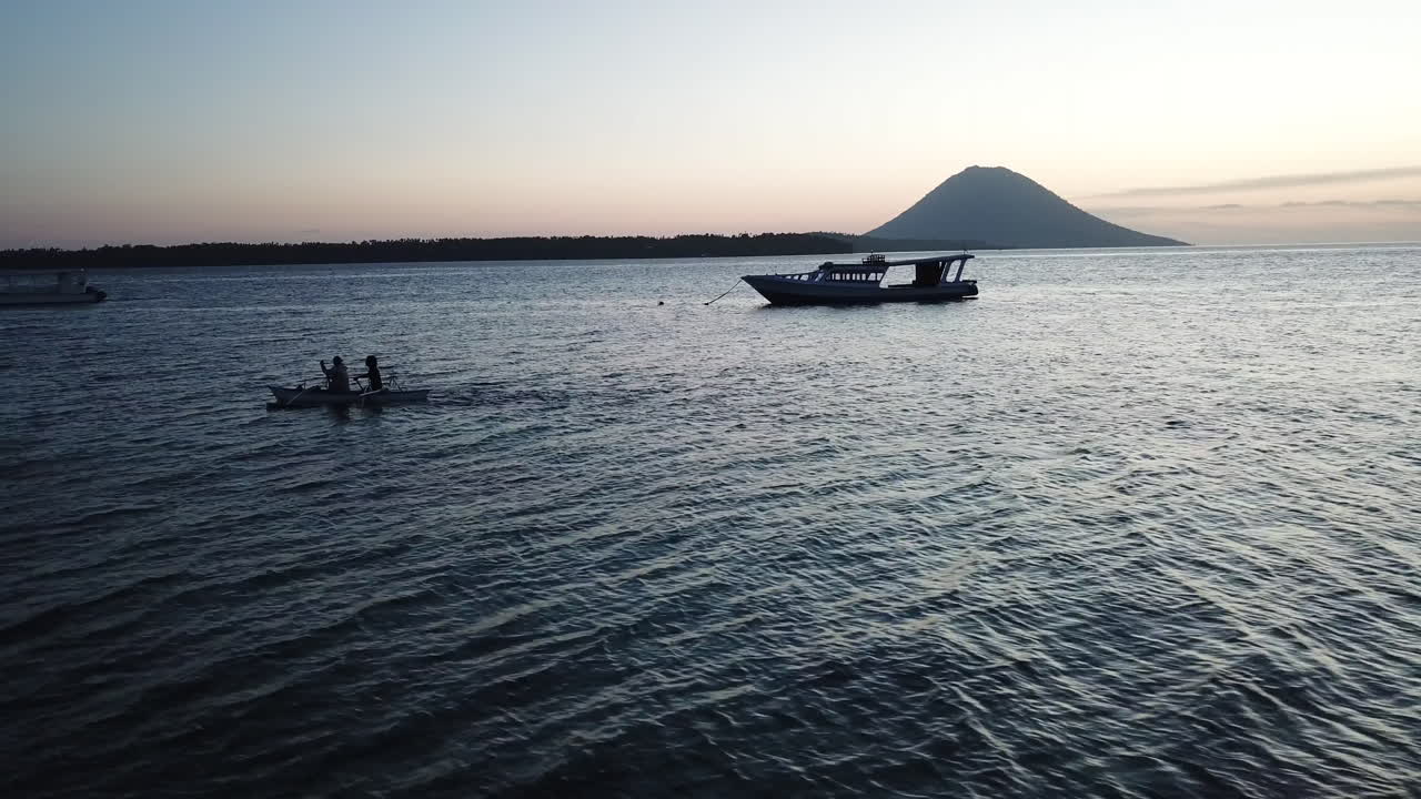 People are kayaking in the ocean during sunset with the silhouette of a boat and volcano in the background.