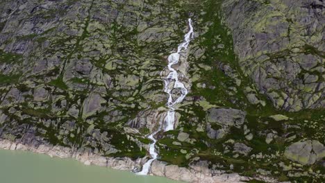 Aerial-drone-zoom-in-shot-over-a-waterfall-along-Grimselsee-close-to-Grimselpass-high-mountain-road-in-beautiful-Swiss-Alps,-Switzerland-on-a-cloudy-day