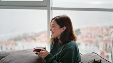 Side-view-of-a-happy-brunette-girl-with-long-hair-in-a-green-sweater-sits-on-a-modern-sofa-with-a-pillow-on-her-feet-and-the-girl-drinks-tea-from-a-brown-glass-in-a-modern-apartment-overlooking-the-sea