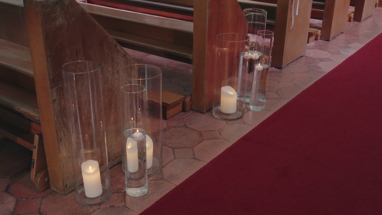 White candles in tall glass vases placed beside red carpet aisle