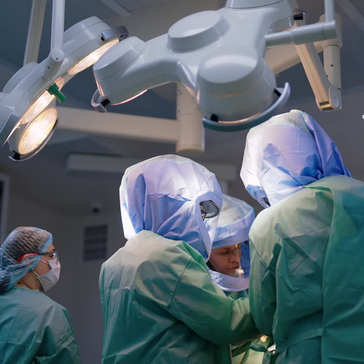 Three doctors work in a surgical team. Professionals wearing protective helmets stand together under the lamps in surgery room