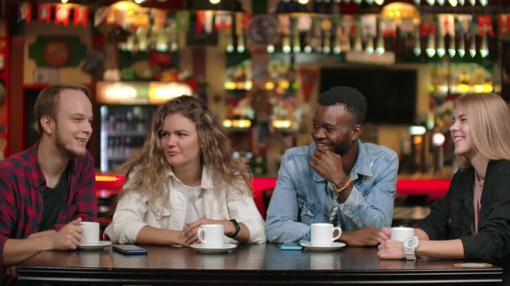 A multinational company of student friends drinks coffee and talks in a restaurant. Four friends chatting laughing and smiling.