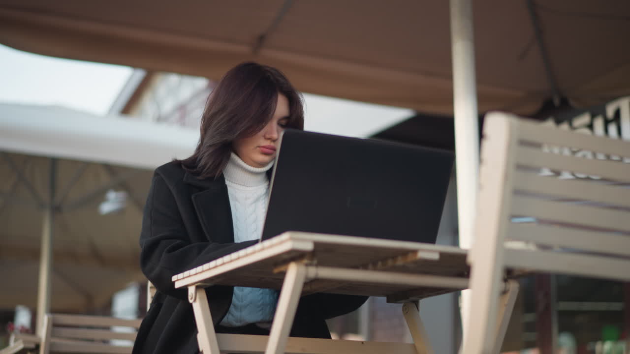 Lower angle view of a woman typing on a laptop while seated outdoors under a patio, dressed in a stylish black coat, with urban scenery and architectural details in the blurred background