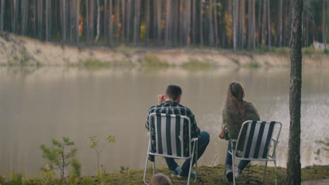 couple of hikers is admiring landscape with lake in forest man and woman are sitting in chair in woodland camp and relaxing