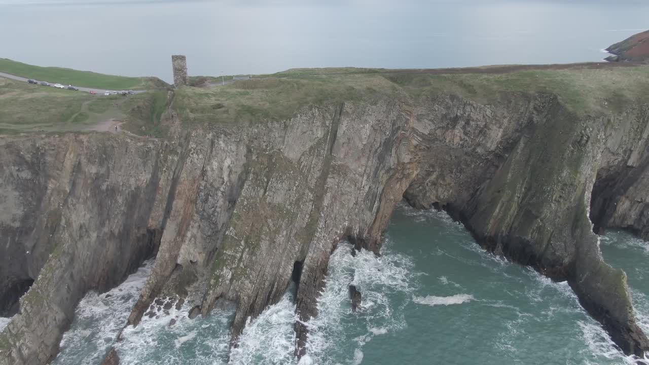 Slow aerial truck right alongside a sea side cliff with rough seas and waves hitting the cliffs and green hills and castle ruin