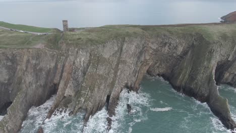 slow aerial truck right alongside a sea side cliff with rough seas and waves hitting the cliffs and green hills and castle ruin