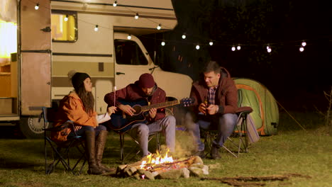 man singing a song on guitar for his friends around camp fire