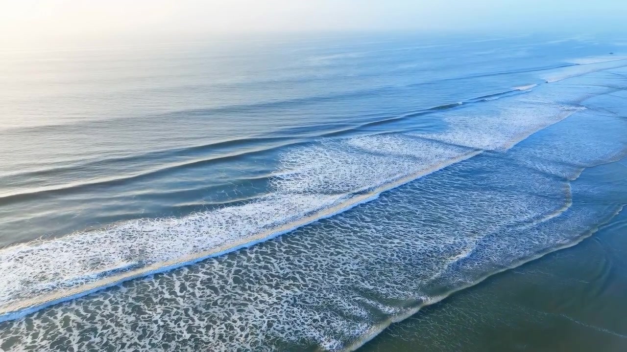 Vista aerea di onde oceaniche che si infrangono sulla costa della spiaggia, mostrando una serena scena naturale con acqua blu e bianca e sabbia rilassanti. Questa tranquilla ambientazione da spiaggia è perfetta.