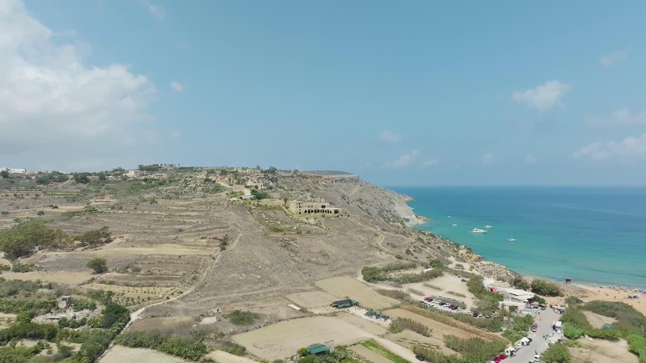 un impresionante panorama aéreo que muestra una pintoresca playa con aguas azules, salpicada de barcos y enmarcada por vegetación natural y afloramientos rocosos y el campo en el fondo