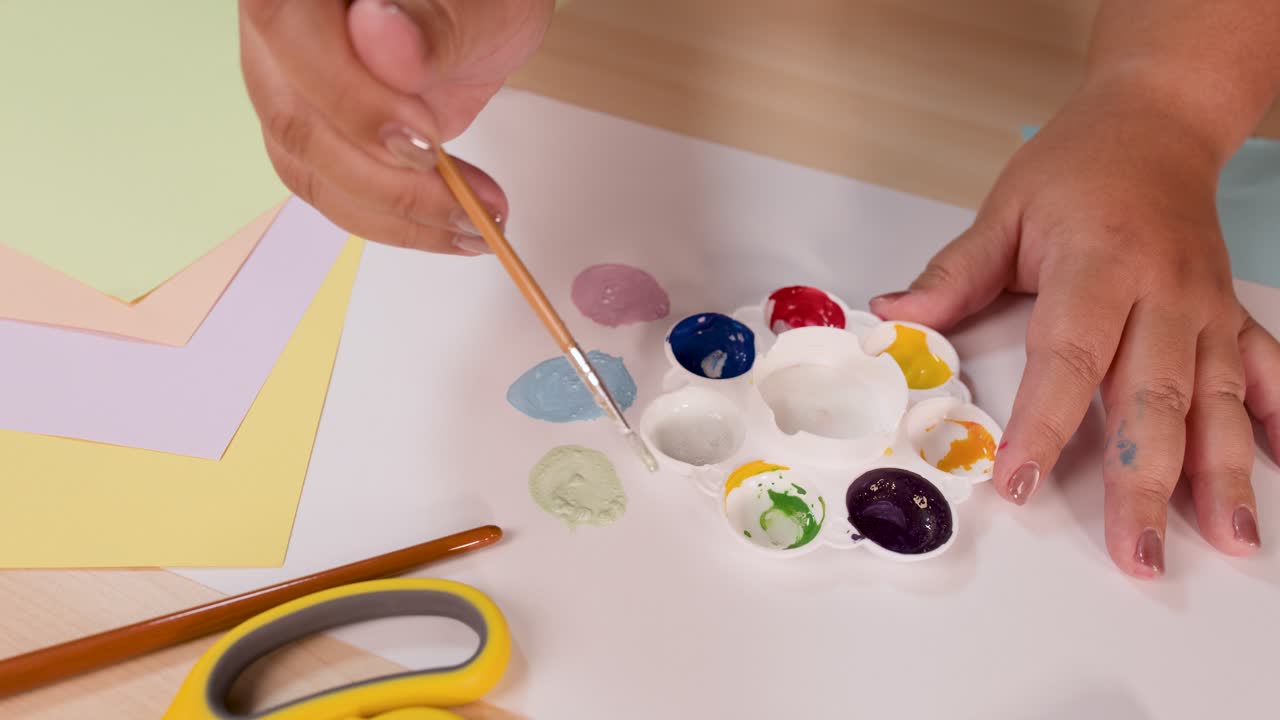 Person mixes watercolor paints with brush on palette, pastel paper background, bright overhead lighting