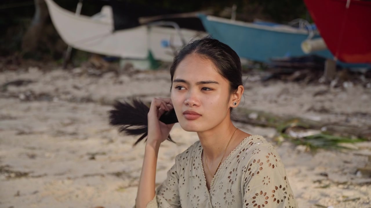 砂浜にいるアジア人女性のクローズアップ。髪には黄色い花を挿し、物思いにふける眼差し。日差しを浴びた肌、背景には停泊するボート。自然な横顔で繊細なドレスをまとい、物思いにふける雰囲気。海辺の静寂と水面の反射。