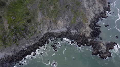 drone overhead shot of waves cascading over the black, rocky coastline of california on a cloudy day in summer. the ocean clarity is contrasted by the green shrubs found on the cliff side above.