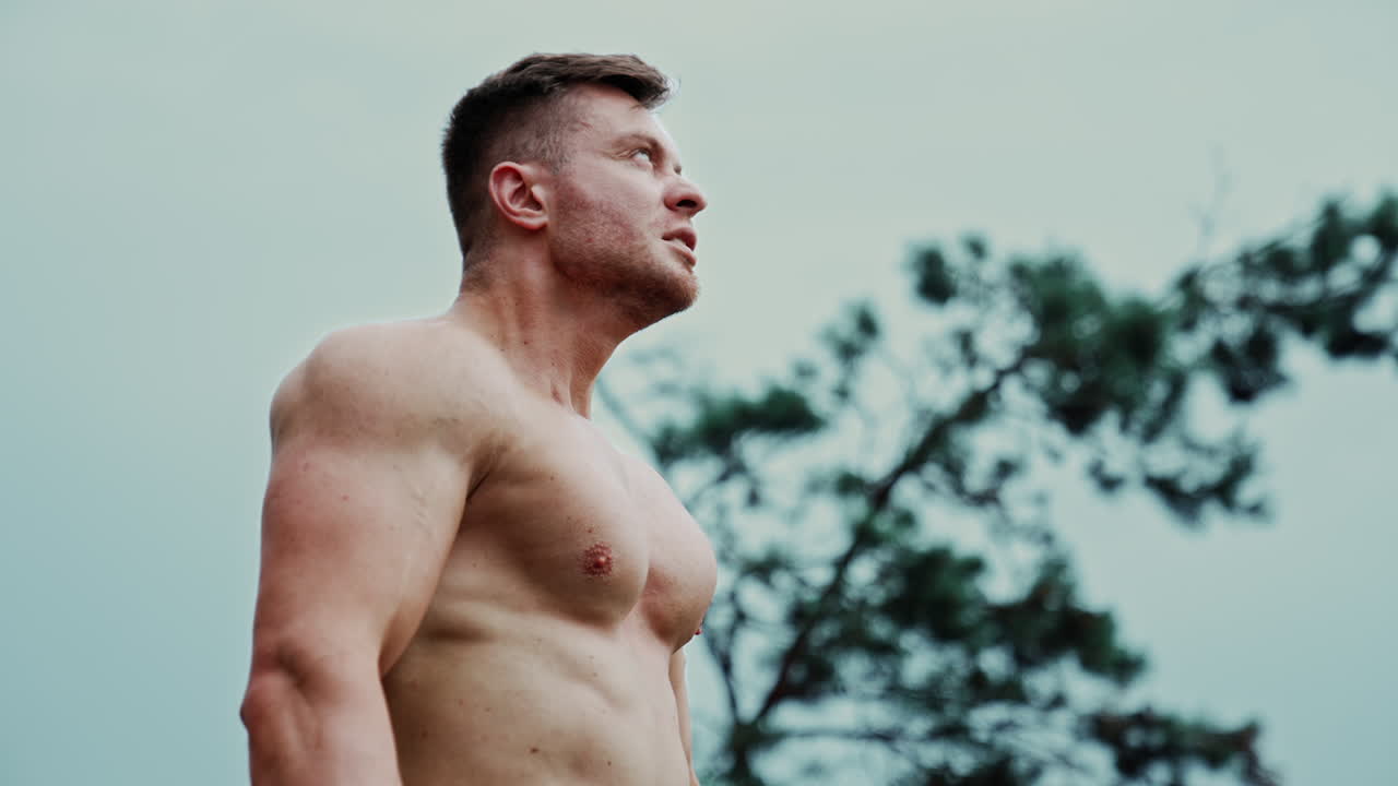 Athletic Caucasian man with naked torso breathing hard after workout. Sportsman exercising outdoors. Low angle view.