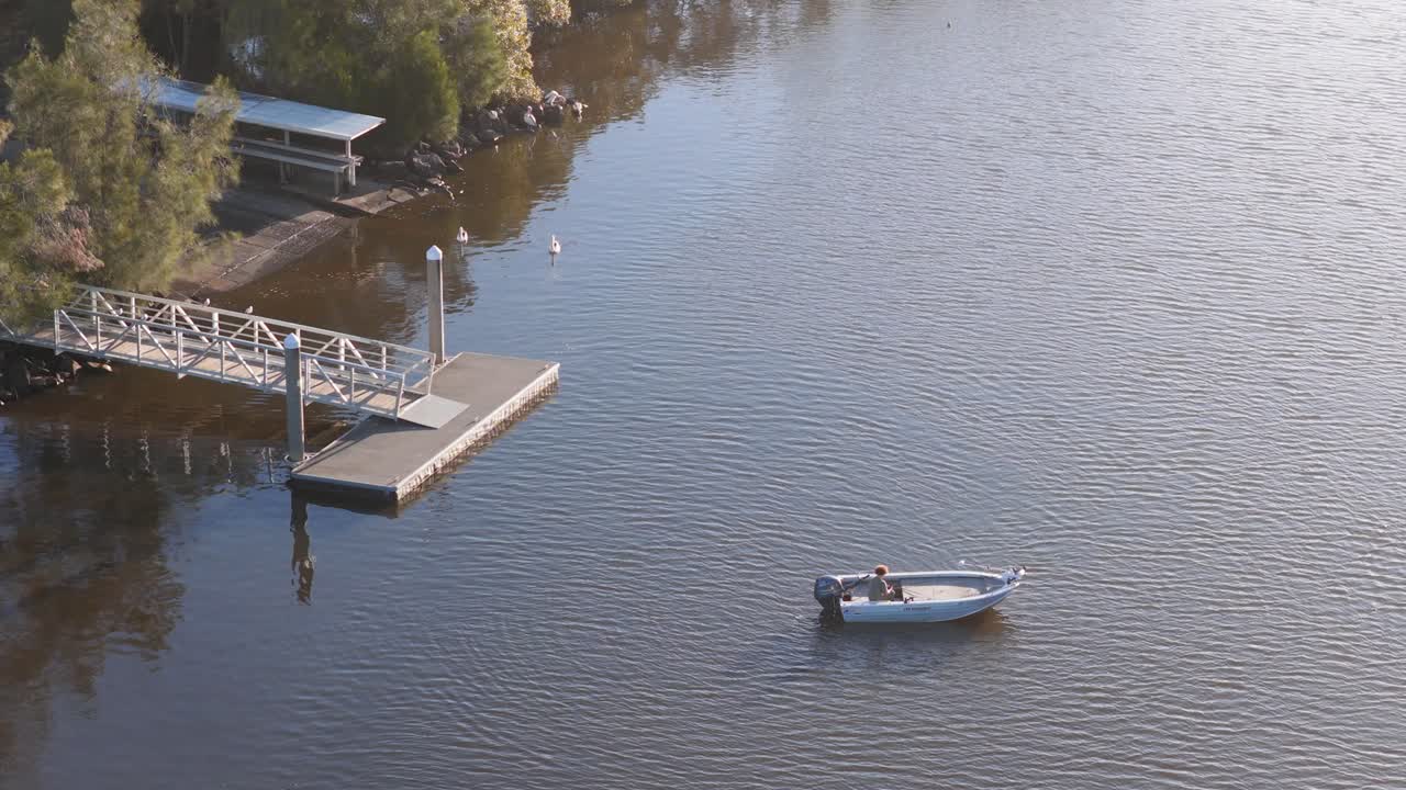 Aerial footage captures a small boat on a calm river near a dock, surrounded by lush greenery under soft lighting