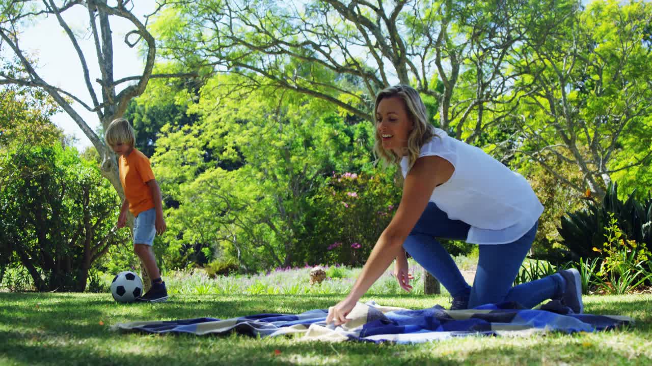madre extendiendo la manta de picnic y hijo jugando al fútbol en el parque 4k