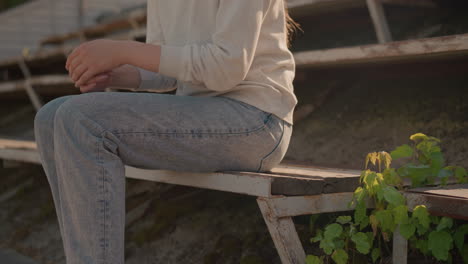 close-up of woman in jeans sitting on first row of rustic stadium seating with aged, rusty metal structure and small green plants growing nearby