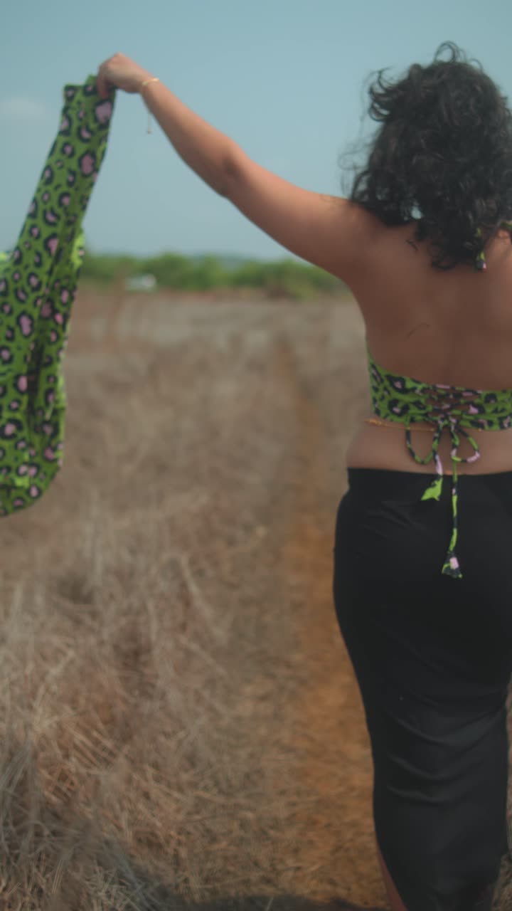 Back view of carefree woman walking outdoors, holding scarf in dry grass field