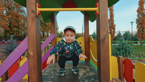 caucasian toddler hangs on the horizontal bar and slides down by the slide. baby boy on the playground in autumn.
