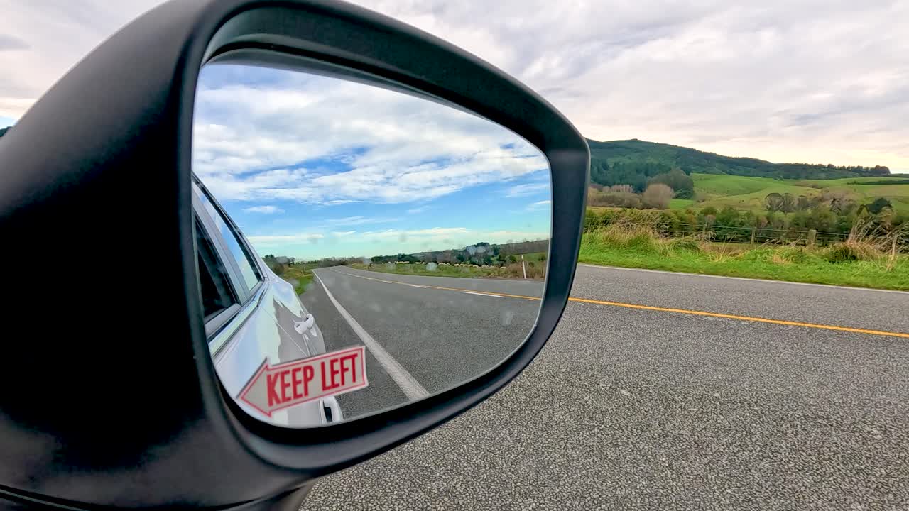 A vehicle overtakes another on a scenic road, captured through a side mirror reflection with clear skies and lush greenery