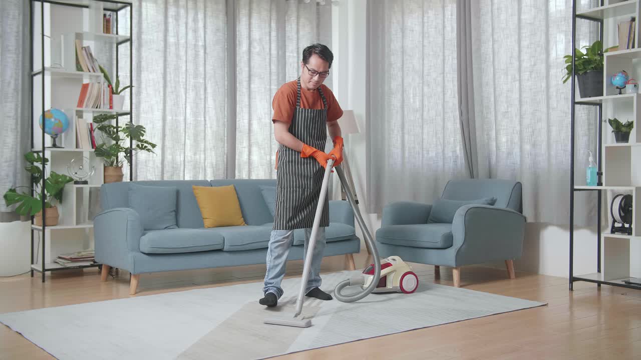 Asian Male Housekeeper With An Apron Enjoys Vacuuming The Floor At Home