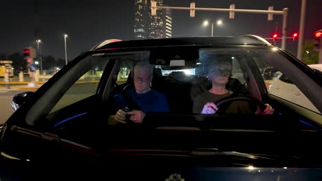 older couple driving through abu dhabi, united arab emirates at nighttime with street decorated for ramadan holiday - view looking into the car through the windshield - husband checking his smartphone