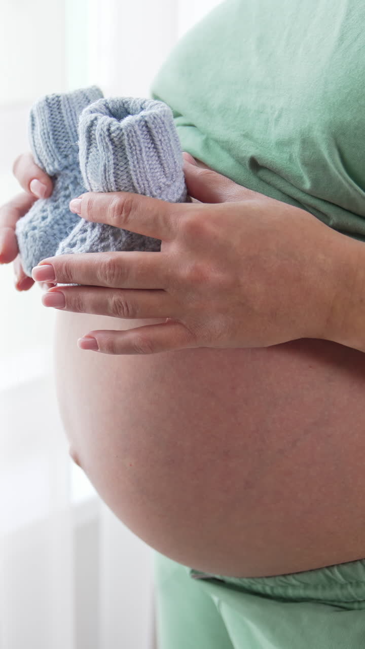 Unidentified pregnant woman paces small baby socks on the top of her big tummy and moves them slowly downwards. Future mom plays with knitted baby booties. Close up. Vertical video