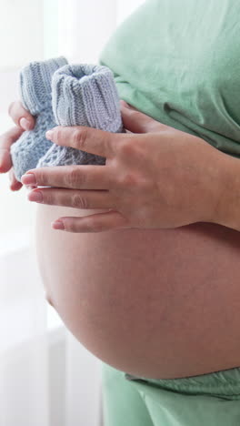 unidentified pregnant woman paces small baby socks on the top of her big tummy and moves them slowly downwards. future mom plays with knitted baby booties. close up. vertical video