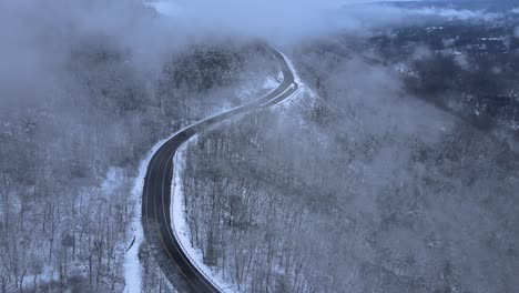 flying through clouds over snow-covered mountains and forests in a mountain valley with a scenic highway below, on a cloud winter’s day