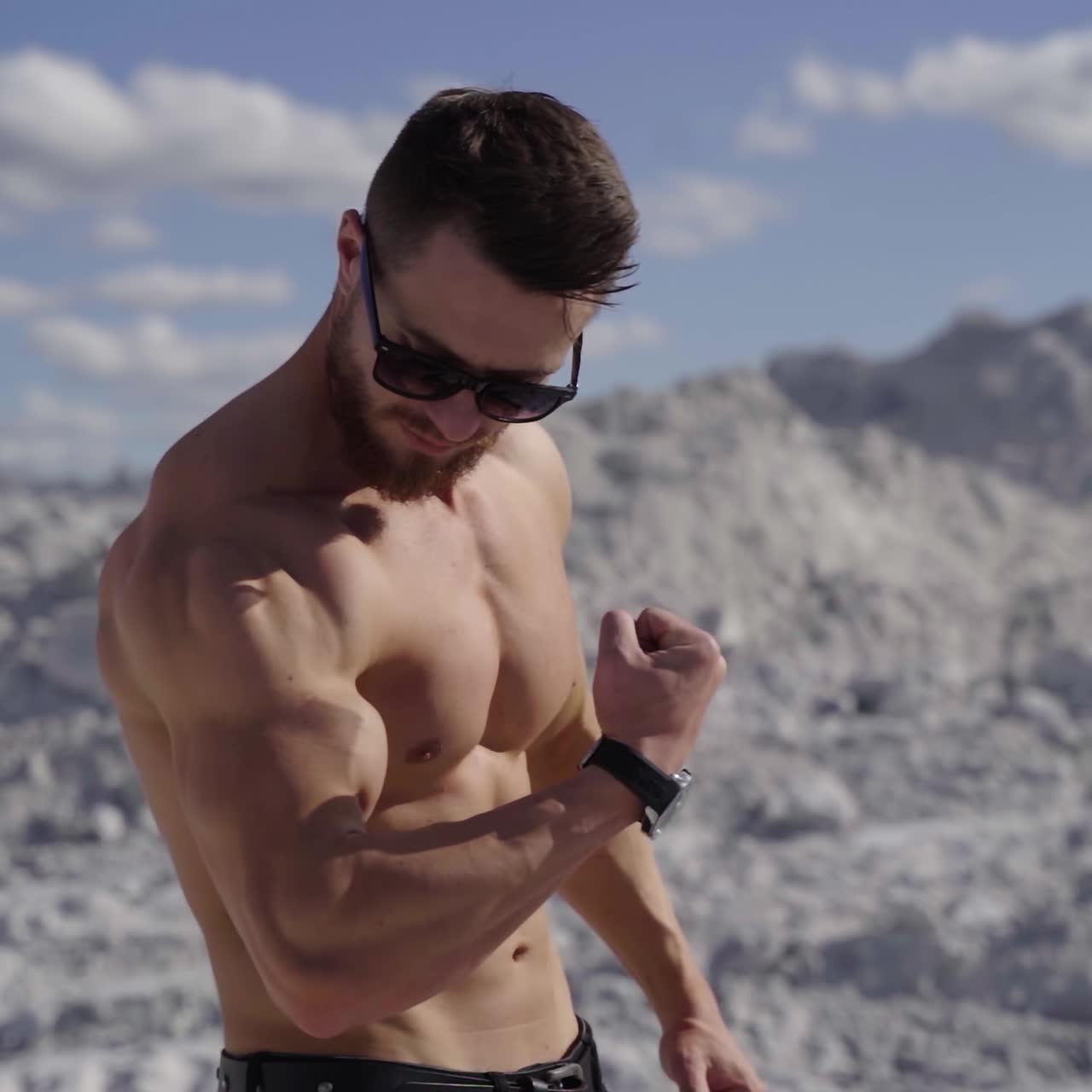 Handsome bodybuilder on the white hill. Strong athlete without shirt shows his arm biceps on nature background. Healthy and sport lifestyle. Slow motion.