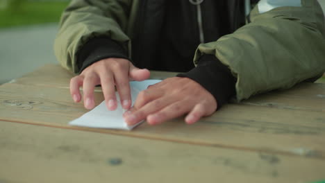 Kid wearing green jacket folds paper kite with focus at outdoor wooden table, hands carefully creasing edges while sitting in park environment on bright day with greenery in soft background