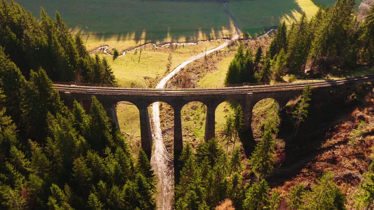 Aerial view of the Telgárt stone railway viaduct in Slovakia, surrounded by dense forest and warm sunlight highlighting the arches and valley path below