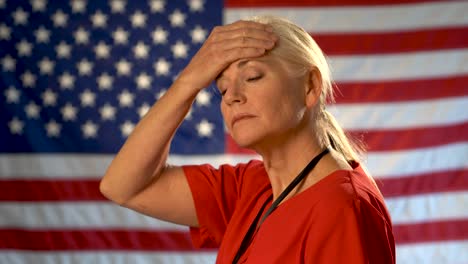 Medium-tight-portrait-of-the-back-of-blonde-nurses-head-as-she-turns-looking-very-worried-and-sad-rubbing-her-forehead-with-American-flag-behind-her