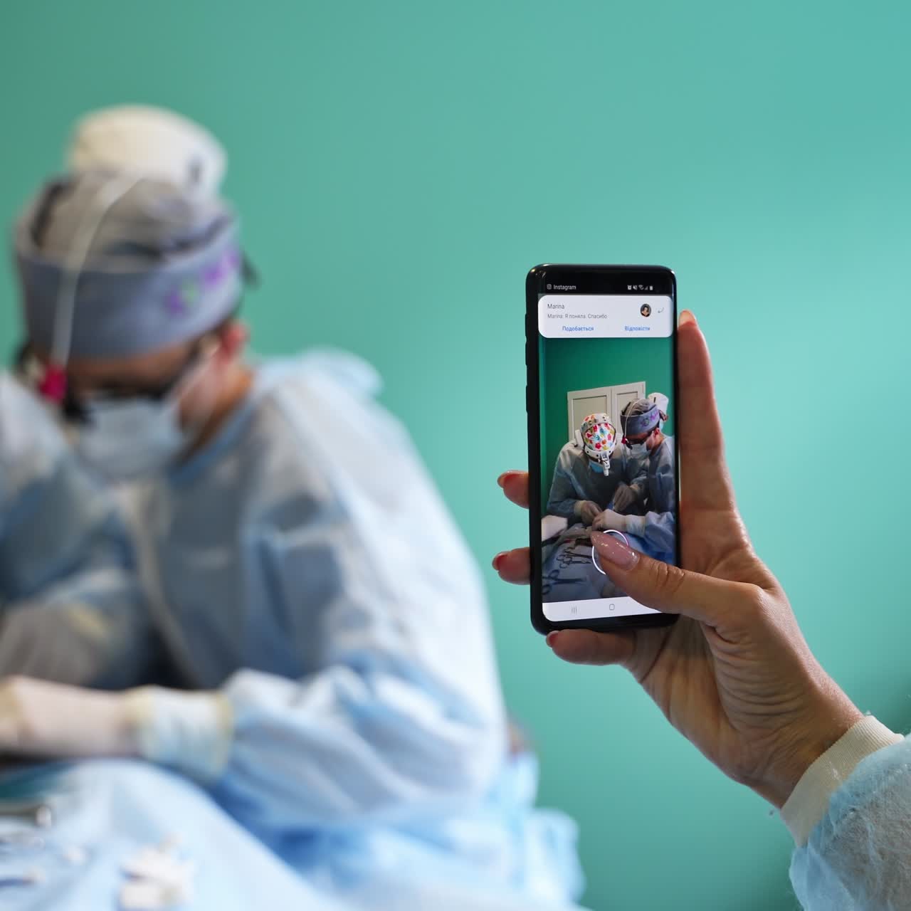 Woman's hand holding a smartphone and taking video of surgery. Two surgeons conducting operation in blur at backdrop