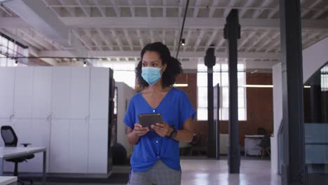 Mixed-race-businesswoman-walking-through-corridor-wearing-facemask-holding-tablet